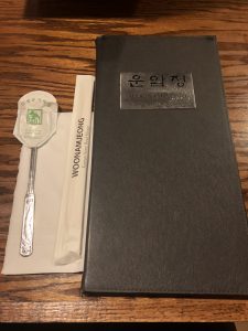 A matte black menu beside a set of cutlery. The menu has a metallic square on the front that says "Stone Bowl House" in English and hangul. 